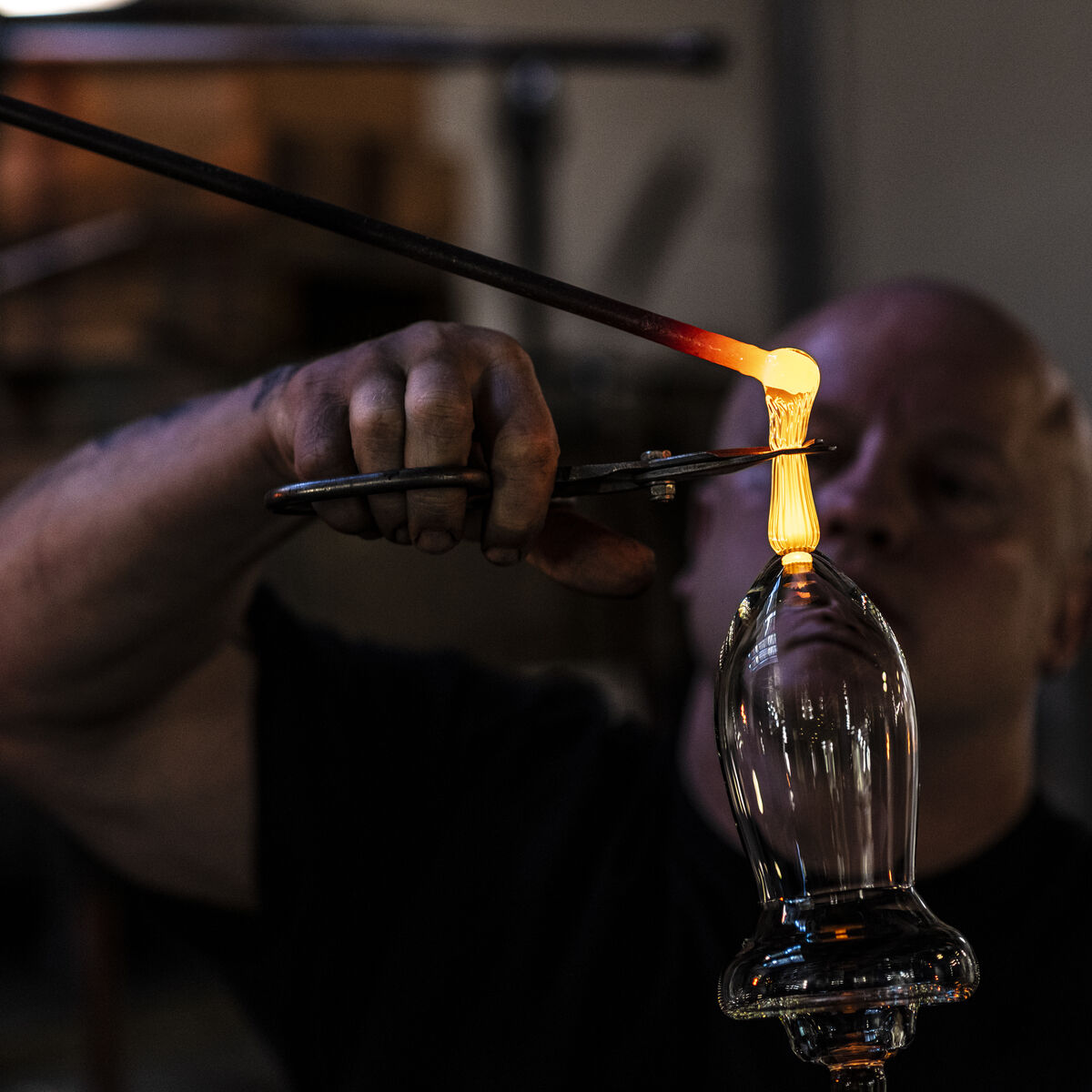 Ein Handwerker stellt mit einer heißen Flamme ein Glas her, indem er es erhitzt und formt. Daneben sind Werkzeuge und weitere Glasarbeiten zu sehen.