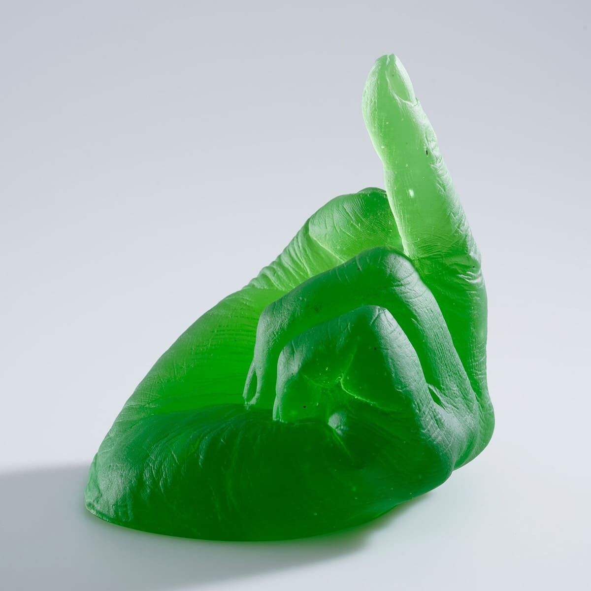 Eine grüne Skulptur in Form einer Hand aus Glas, die den Mittelfinger hochhält, entworfen von Ai Weiwei. Die Skulptur ist in einer transparenten, satten Grüntönung gehalten und steht vor einem neutralen Hintergrund.