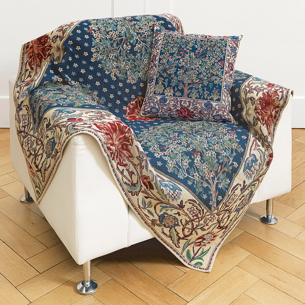 Eine dekorative Kissenhülle mit dem Motiv 'Tree of Life' nach William Morris liegt auf einem weißen Sessel, daneben eine farblich passende Tagesdecke. Das Design zeigt ein reich verziertes florales Muster mit Pflanzen und Vögeln auf einem blauen Hintergrund. Das Produkt befindet sich in einem hellen Wohnzimmer mit Parkettboden.