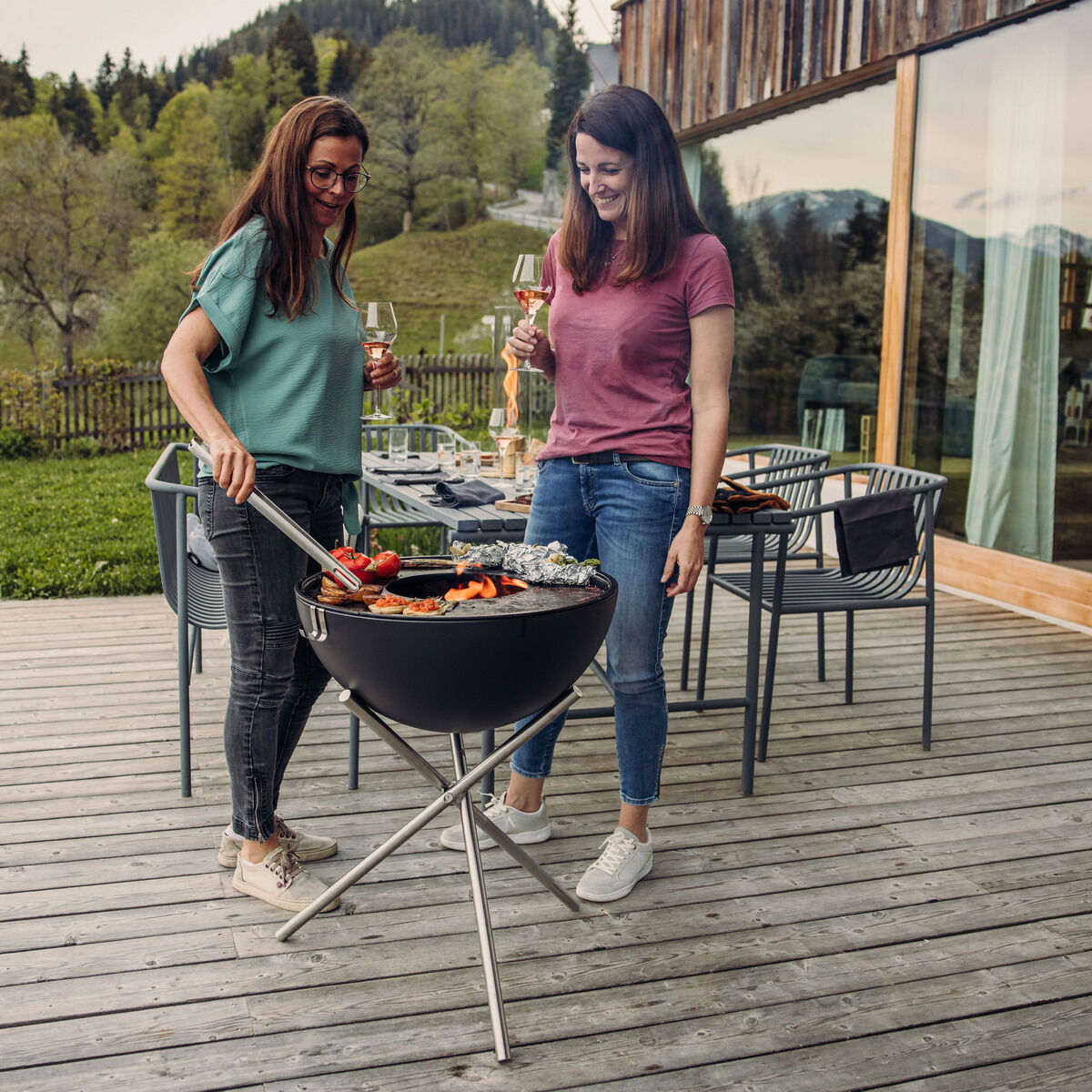 Zwei Frauen grillen auf einem Plancha-Grill auf einer Außenterrasse. Sie trinken Wein und lächeln, während sie Gemüse und Fleisch grillen. Im Hintergrund sind grüne Bäume und ein Holzhaus zu sehen.