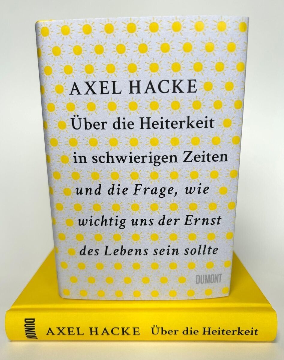 Das Bild zeigt das Buch 'Über die Heiterkeit in schwierigen Zeiten' von Axel Hacke. Das Buch liegt auf einem gelben Buchdeckel, und der Einband des Buches ist weiß mit gelben Punkten und schwarzem Text.