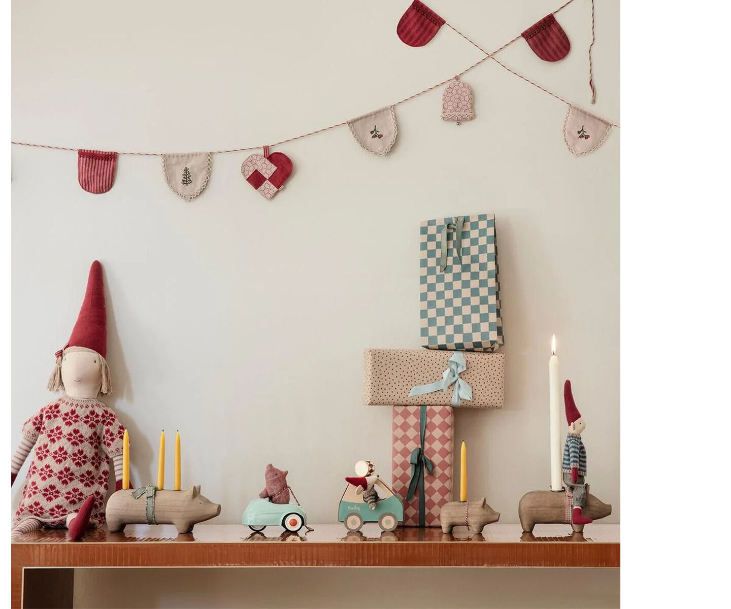 Auf einem Regal stehen mehrere Maileg-Figuren, darunter die Weihnachtsmaus im grünen Auto mit roter Zipfelmütze und Strickpullover, umgeben von Tieren mit Kerzen und weihnachtlicher Deko. Darüber hängen festliche Girlanden; im Hintergrund bunte Geschenke.