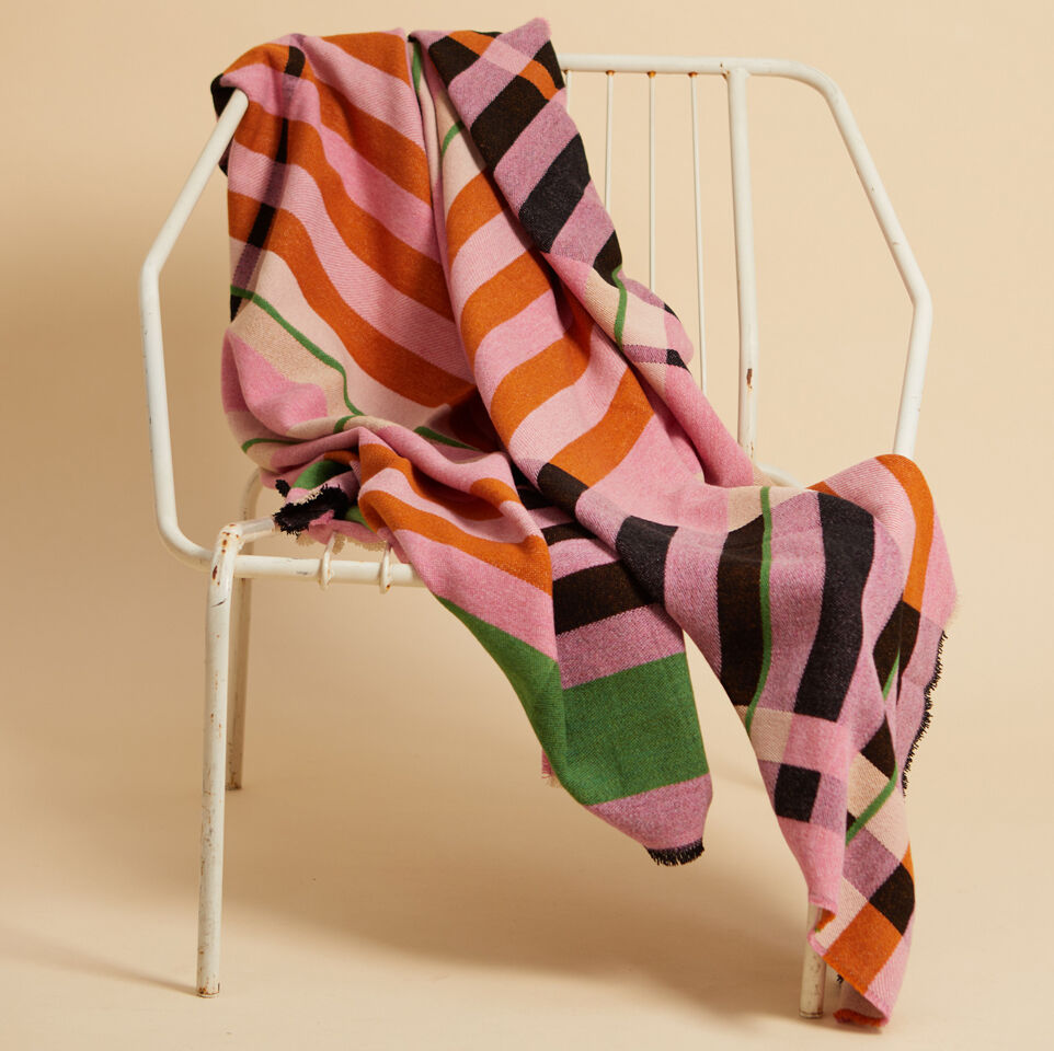 Eine Decke mit buntem Streifenmuster, darunter Rosa, Orange, Schwarz, Grün und Lila Farben, drapiert über einen weißen Stuhl. Der Hintergrund ist beige.