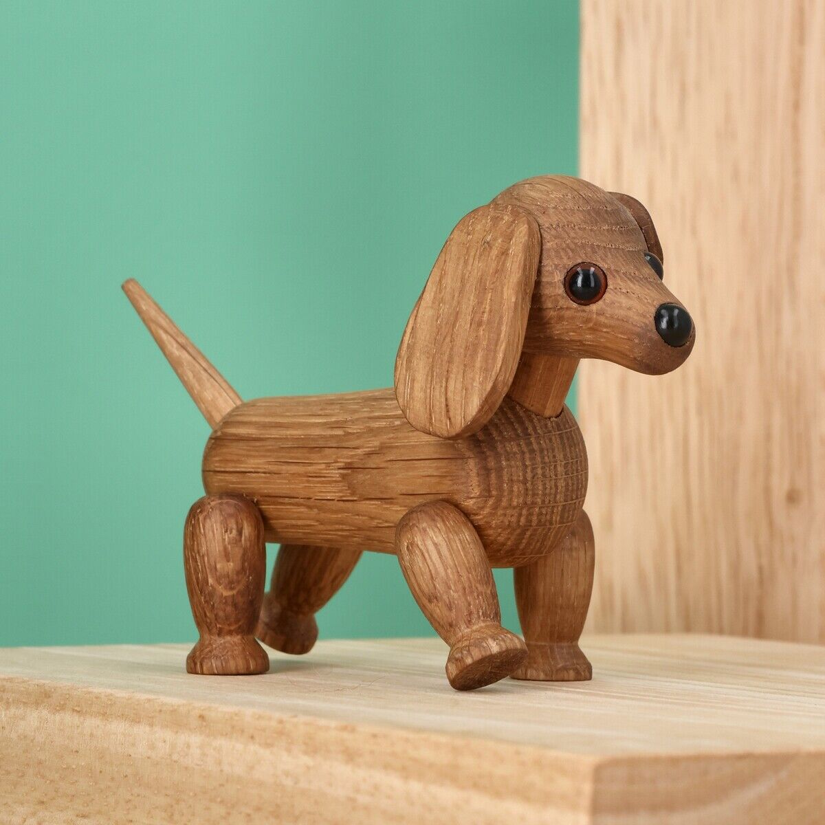Detaillierte Holzfigur eines Dackels mit dunklen Knopfaugen, langen Ohren und abstehendem Schwanz. Die Figur steht auf einem hellen Holzpodest vor einer mintgrünen und einer hellen Holzwand. Produkt ist die Holzfigur »Dackel Chico« von Spring Copenhagen.