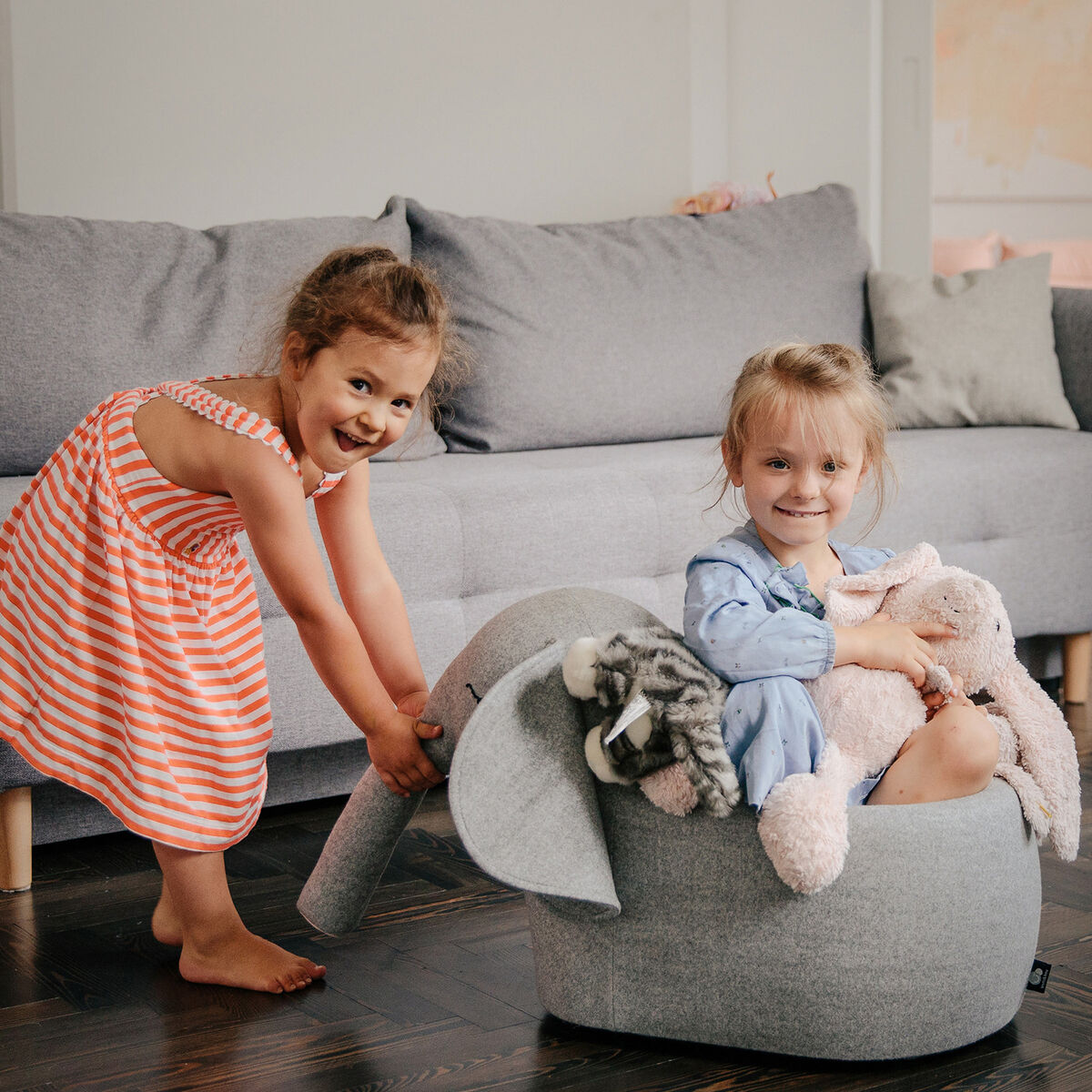 Zwei lächelnde Mädchen spielen im Wohnzimmer mit einem Rollelefant und Kuscheltieren.