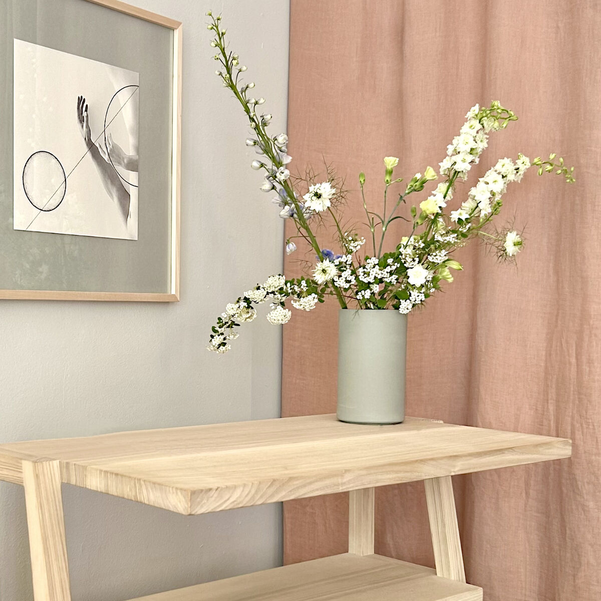 Eine minimalistische Szene zeigt den Weinkühler und Vase "KULA" in betongrün matt von Frau Caze, gefüllt mit weißen und grünen Frühlingsblumen auf einem hellen Holzregal. Dahinter ein gerahmtes abstraktes Bild und ein pastellfarbener Vorhang an der Wand.