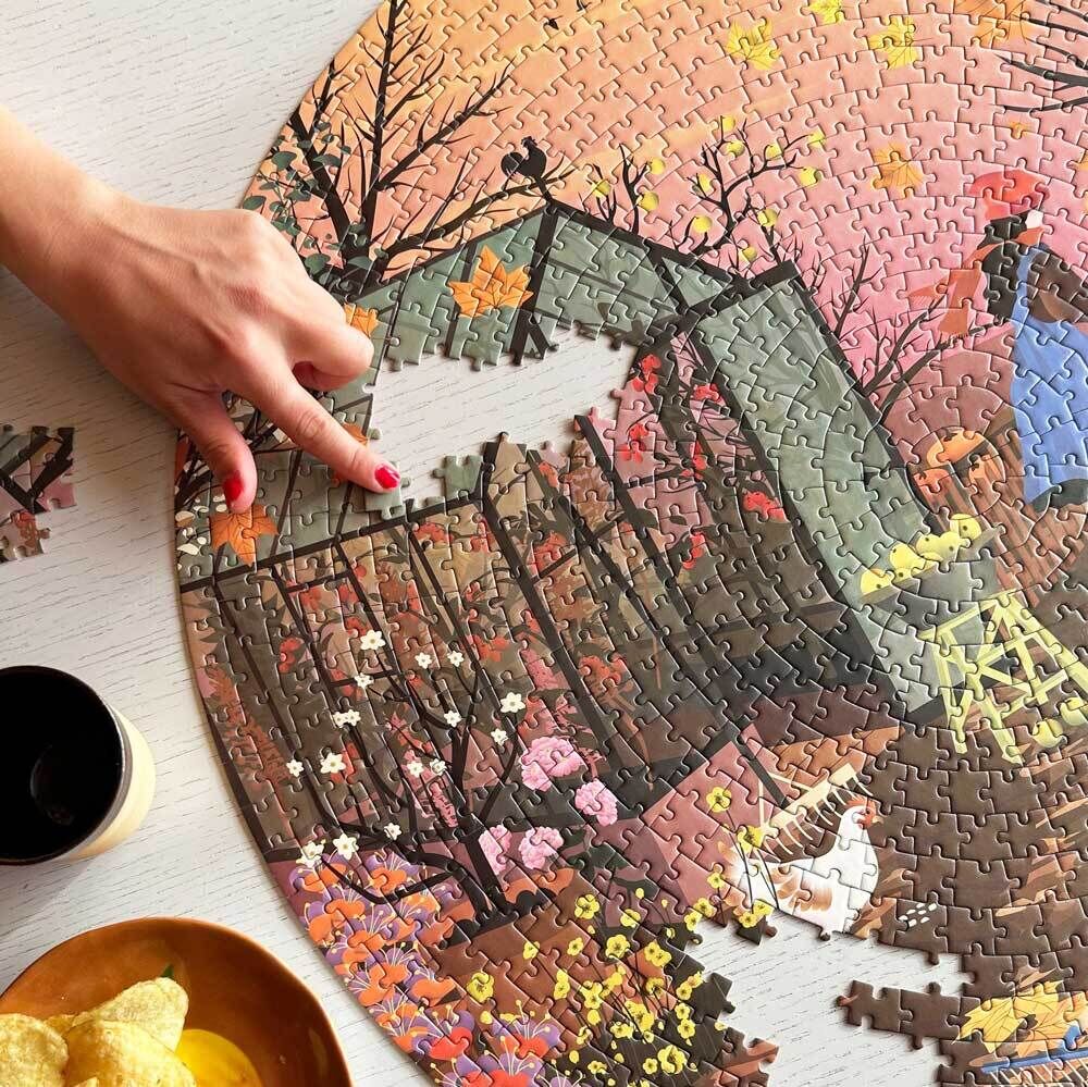 Person fügt ein rundes Herbstgefühle-Puzzle zusammen, das herbstliche Naturmotive zeigt. Im Vordergrund befinden sich eine Schüssel mit Chips und eine Tasse. Die Szene ist auf einem Tisch arrangiert.