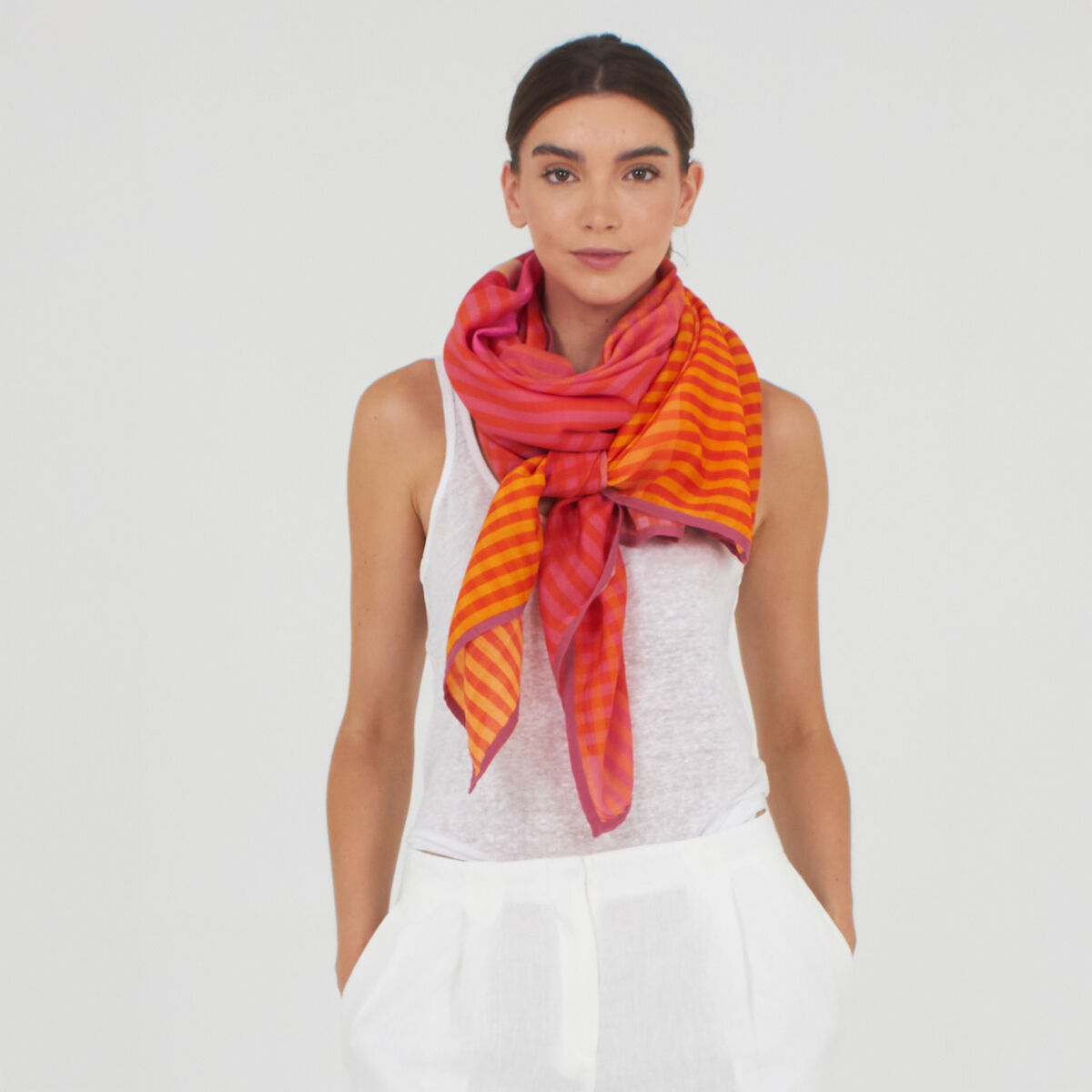 Eine Frau trägt einen großformatigen Baumwoll-Seidenschal »Willa« von Shalls Barcelona um den Hals, auffallend in orange, pink und rot gestreift. Sie trägt ein weißes ärmelloses Top und weiße Hose vor neutralem Hintergrund. Der Schal wirkt leicht und edel.