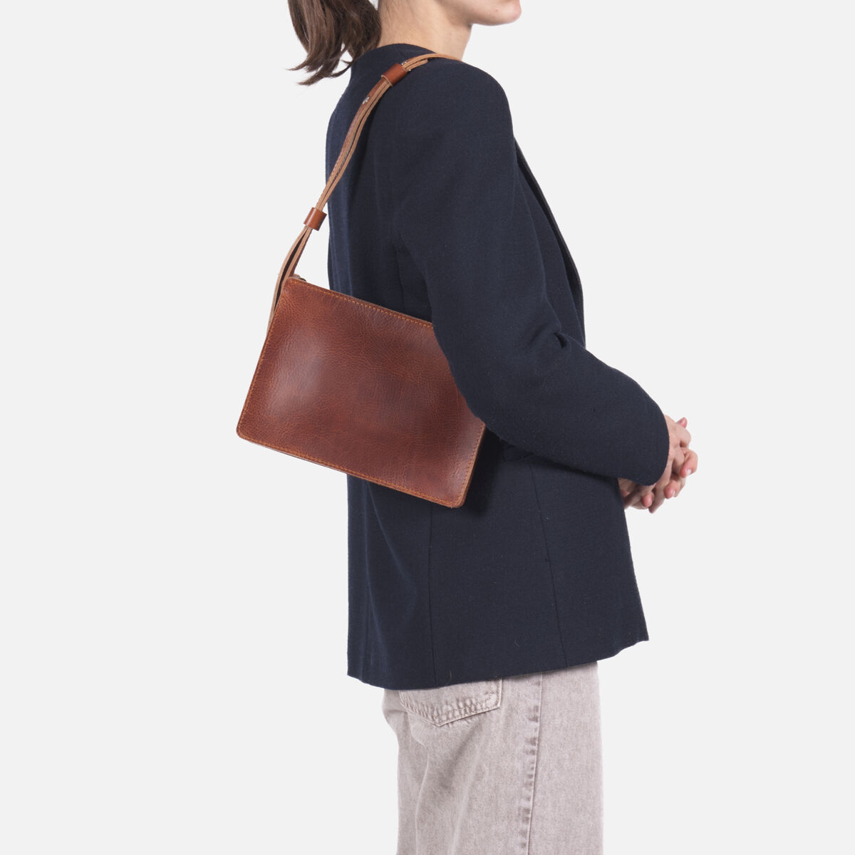 Eine Frau trägt eine schlichte Umhängetasche aus braunem Kernleder über der Schulter. Sie trägt einen dunkelblauen Blazer und helle Jeans. Die Ledertasche wirkt hochwertig, minimalistisch und edel, passend für Alltag oder Business.