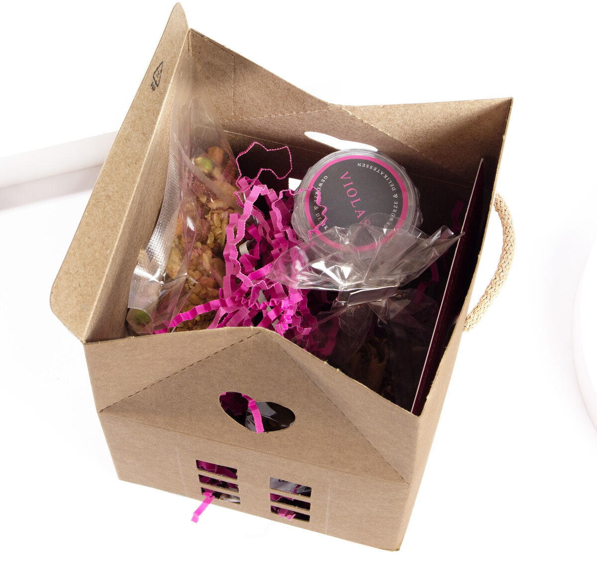 Das Bild zeigt ein Geschenkset in Form eines kleinen Weihnachts-Fachwerkhauses. Die Verpackung ist aus braunem Karton mit einem Sichtfenster auf der Vorderseite und einem Griff oben. Im Inneren finden sich allerlei Süßigkeiten und eine Nussmischung, die mit rosa Papierschnipseln dekorativ verpackt sind.