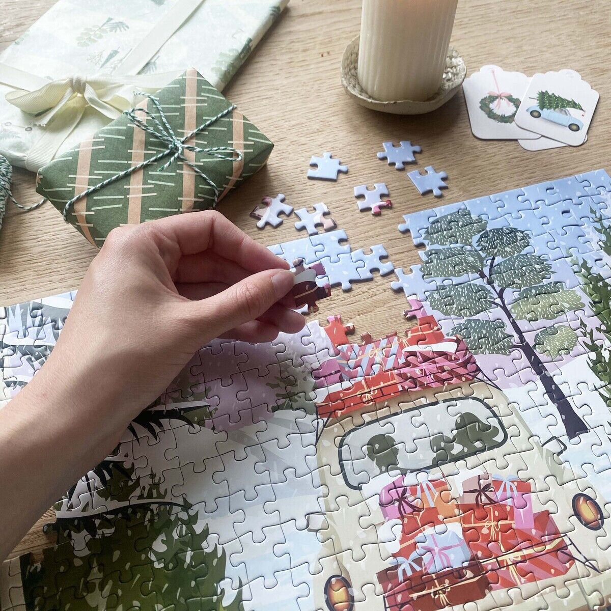 Auf einem Holztisch wird das 300-Teile-Puzzle „Alles gepackt“ der Marke VISSEVASSE zusammengesetzt. Zu sehen ist ein Wohnmobil mit vielen Geschenken auf dem Dach in Landschaft. Daneben stehen verpackte Geschenke, eine brennende Kerze und zwei Geschenkanhänger.