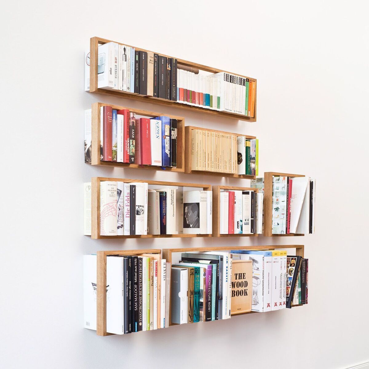 An der Wand montierte Regale aus Holz, die mehrere Bücherreihen in unterschiedlichen Farben und Größen enthalten. Das Regal hat ein minimalistisches und modernes Design.