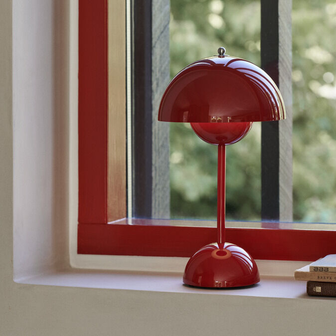Rote, glänzende Flowerpot VP9 Tischleuchte von &Tradition steht auf einer Fensterbank mit rotem Rahmen. Im Hintergrund ist durch das Fenster unscharfes grünes Blattwerk sichtbar. Rechts liegen Bücher auf der Fensterbank. Die Lampe ist klassisch rund und retro designt.
