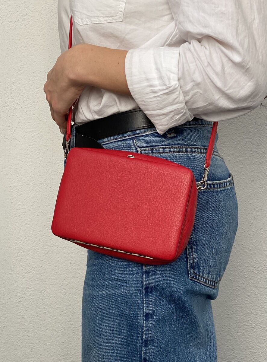Eine Frau trägt eine rote Lederhandtasche mit Schulterriemen. Die Tasche ist klein und hat eine rechteckige Form. Sie trägt eine weiße Bluse und blaue Jeans. Der Hintergrund ist eine weiße Wand.