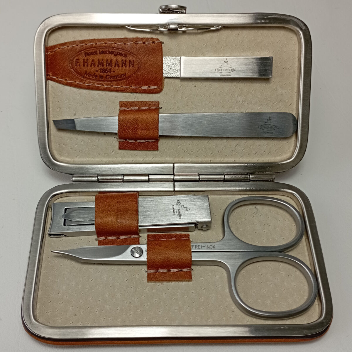 Ein Maniküre-Set in einem eleganten Lederetui. Das Set enthält eine Nagelschere, einen Nagelknipser, eine Nagelfeile und eine Pinzette. Die Werkzeuge sind aus hochwertigem Metall, und das Etui ist in Brauntönen mit einer cremefarbenen Innenausstattung gehalten.