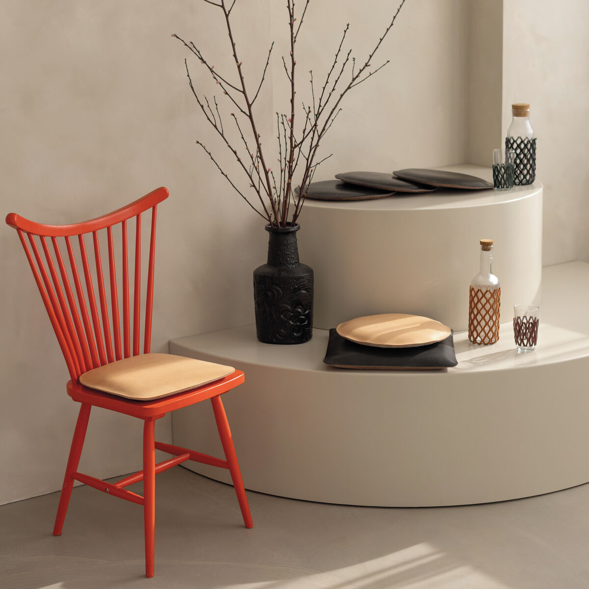 Sitzpolster Rund in Cognac-Farbe von Harold's Lederwaren auf modernem roten Stuhl und runder, weißer Podestfläche. Daneben Deko mit schwarzen und grünen Flaschen, Zweigen in einer schwarzen Vase sowie minimalistischem, hellem Interieur.