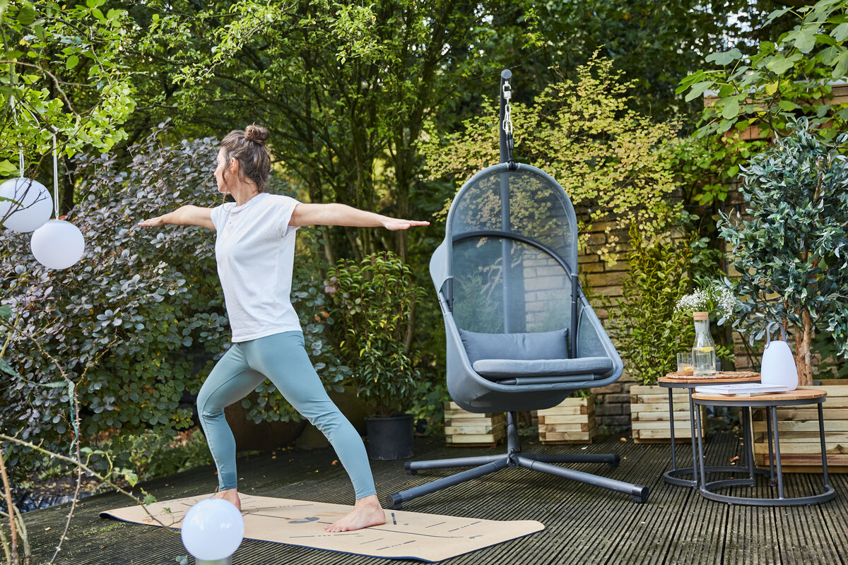 Eine Frau in Yoga-Position auf einer Terrasse neben einem modernen grauen Hängesessel »Yobaya«, umgeben von grünen Pflanzen und Gartenmöbeln.