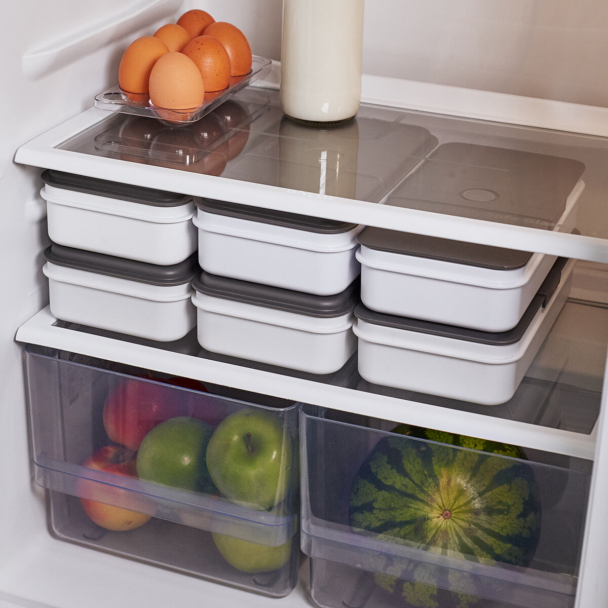 Eine gut organisierte Kühlschrankansicht mit mehreren gestapelten weißen und grauen Lebensmittelbehältern, einem Karton Eier, einer Flasche Milch und frischem Obst, einschließlich Äpfeln, einem Wassermelonenstück und anderen Früchten, in transparenten Schubladen.
