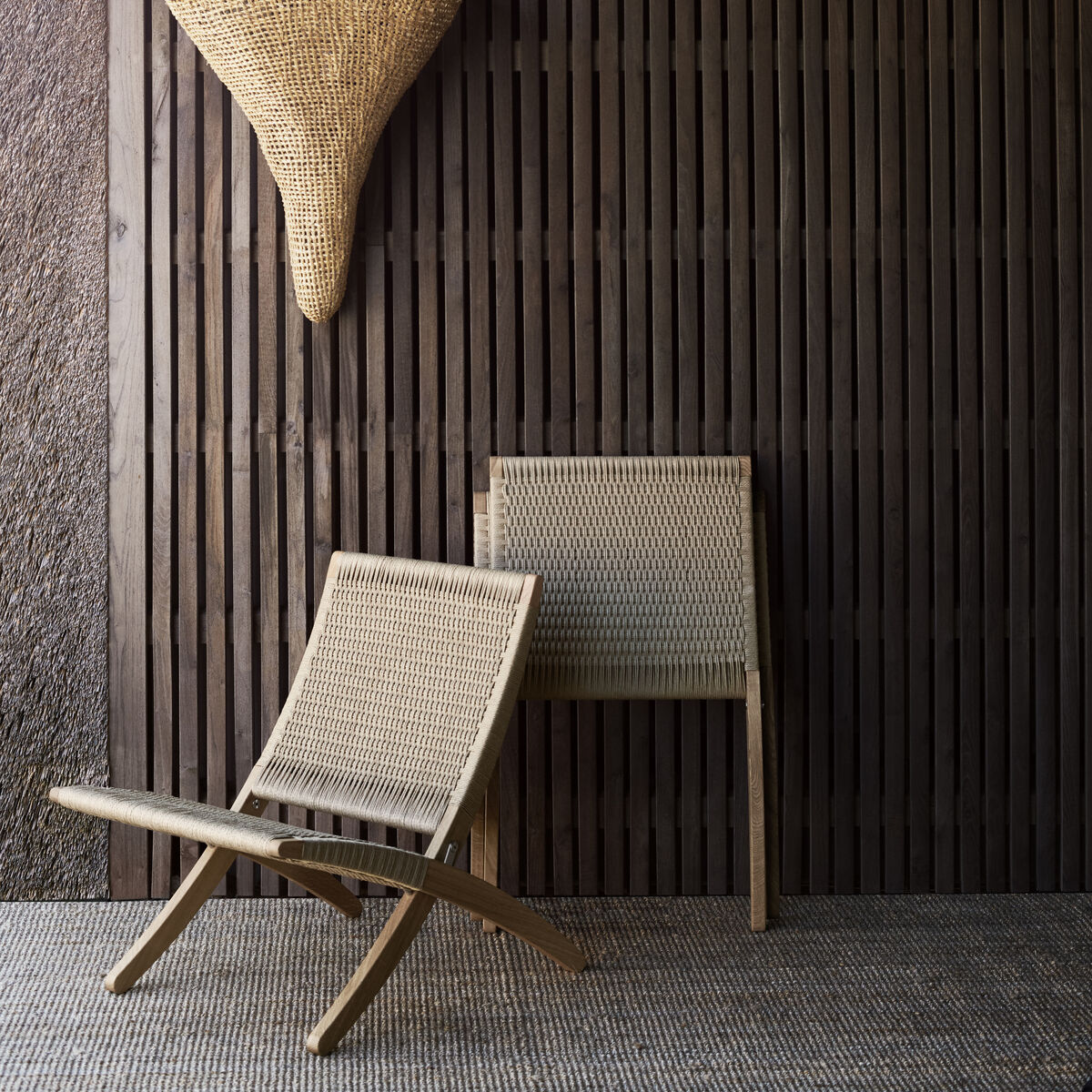 Zwei minimalistische Rattan-Stühle, die auf einem stilvollen, dunklen Holz-Hintergrund stehen. Ein gewebter Wandkorb hängt oberhalb der Stühle. Der Raum wirkt modern und naturbelassen.