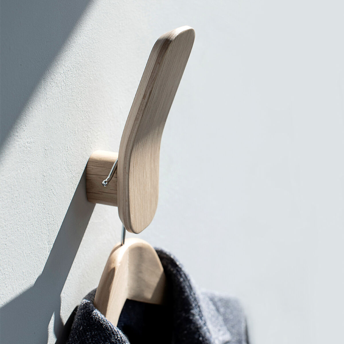 Ein minimalistischer Kleiderhaken aus Eichenholz ist an einer grauen Wand befestigt. An ihm hängt ein graues Kleidungsstück auf einem Holzbügel. Natürliches Licht fällt auf den Haken und die Wand, was dem Bild eine helle, einladende Atmosphäre verleiht.