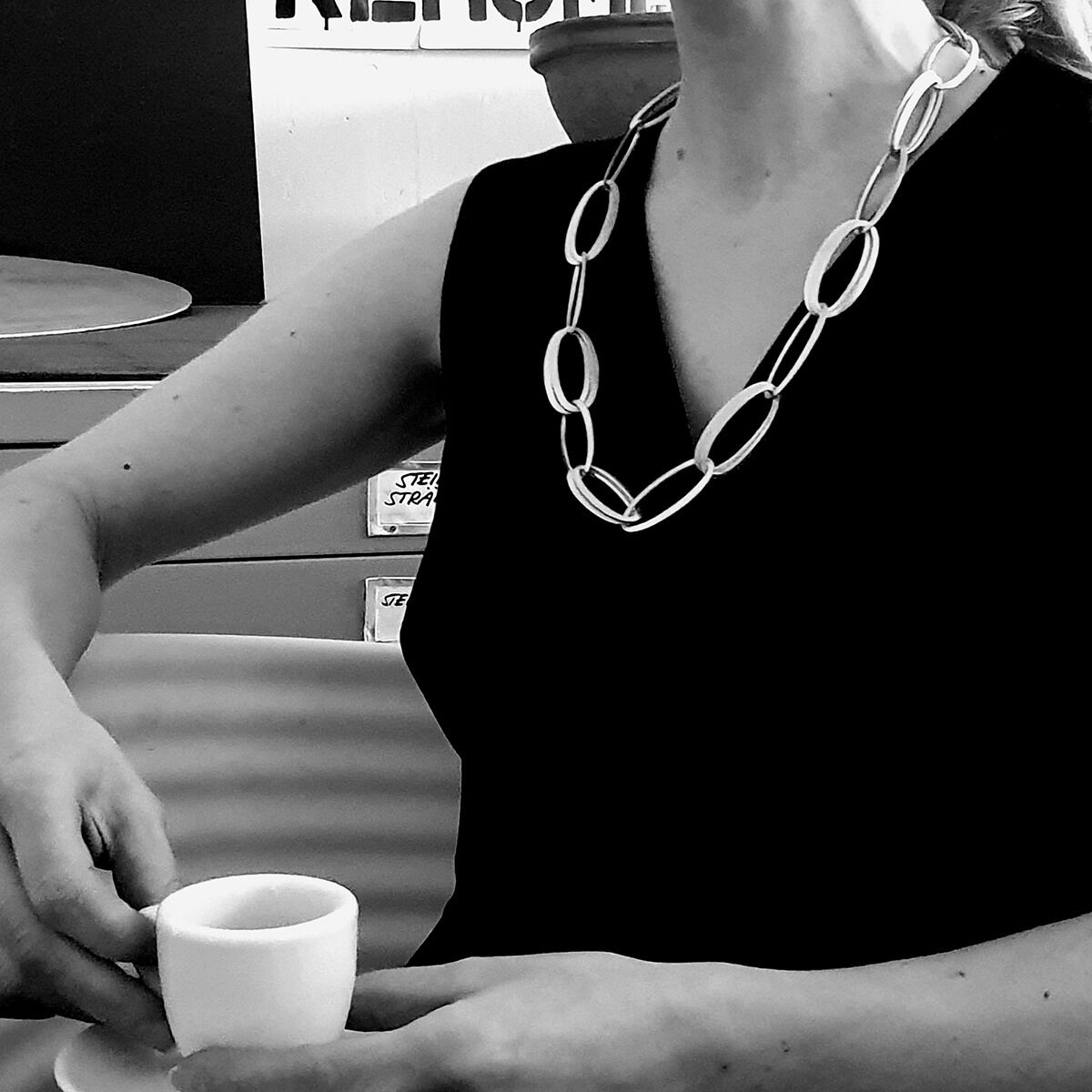 Frau mit schwarzem Oberteil trägt das exklusive Collier »GRACE« von HML Berlin aus ovalen, handgefertigten, silbernen Gliedern. Sie sitzt an einem Tisch mit Espressotasse. Das Schmuckstück wirkt zeitlos und elegant, Foto ist in Schwarz-Weiß gehalten.