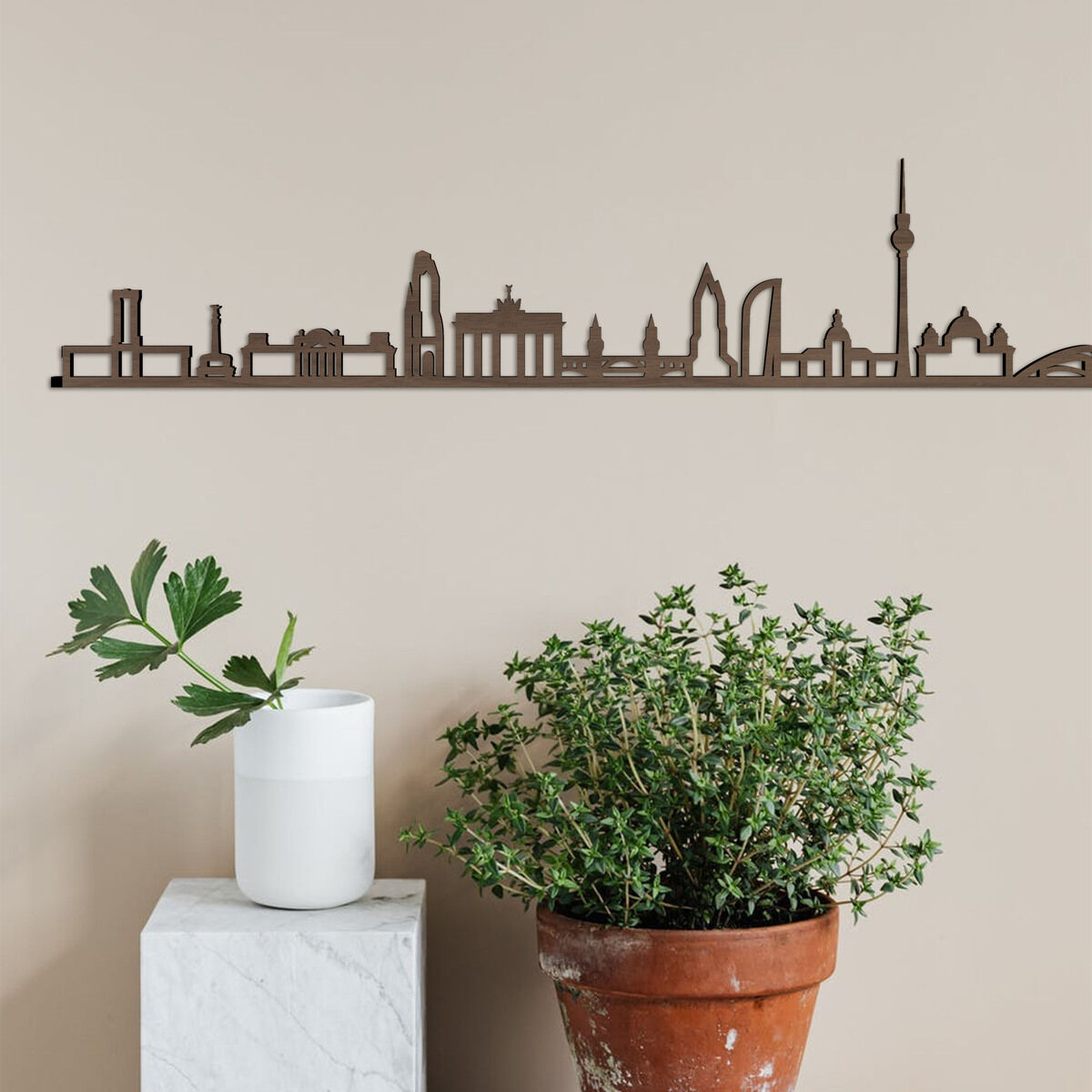 Eine Holzschnitt-Skyline von Berlin hängt an einer beigen Wand. Neben der Skulptur stehen links eine weiße Vase mit grünen Pflanzen auf einem weißen Sockel und rechts ein großer Topf mit einer Pflanze.