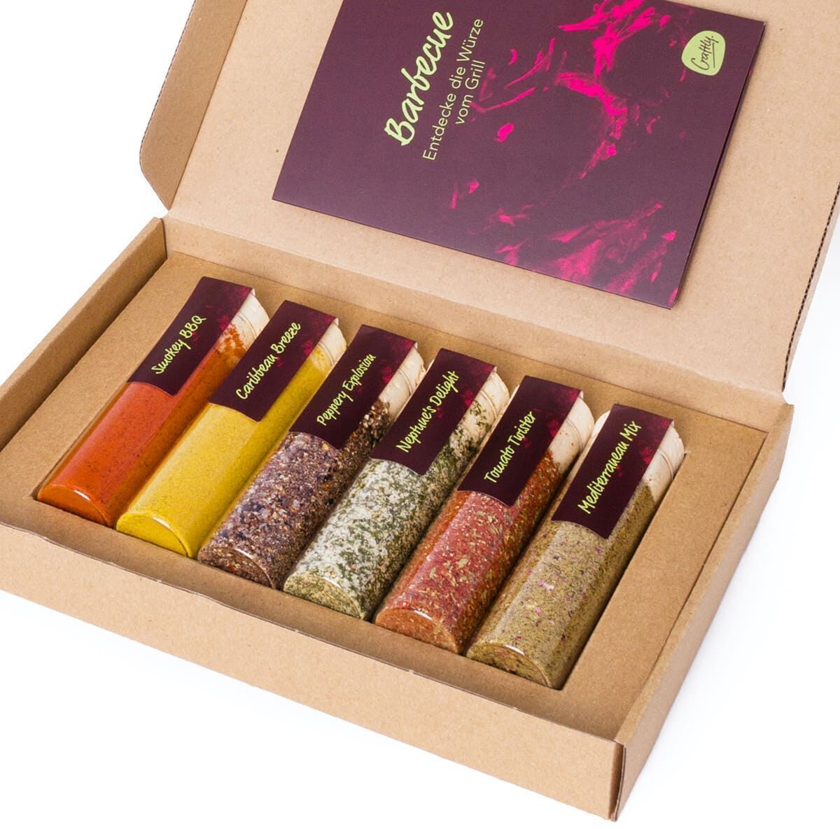 Eine Geschenkbox mit verschiedenen BBQ-Gewürzen in durchsichtigen Röhrchen, die ordentlich in einer braunen Pappbox angeordnet sind. Auf der Innenseite des Deckels befindet sich ein lila-farbenes Etikett.