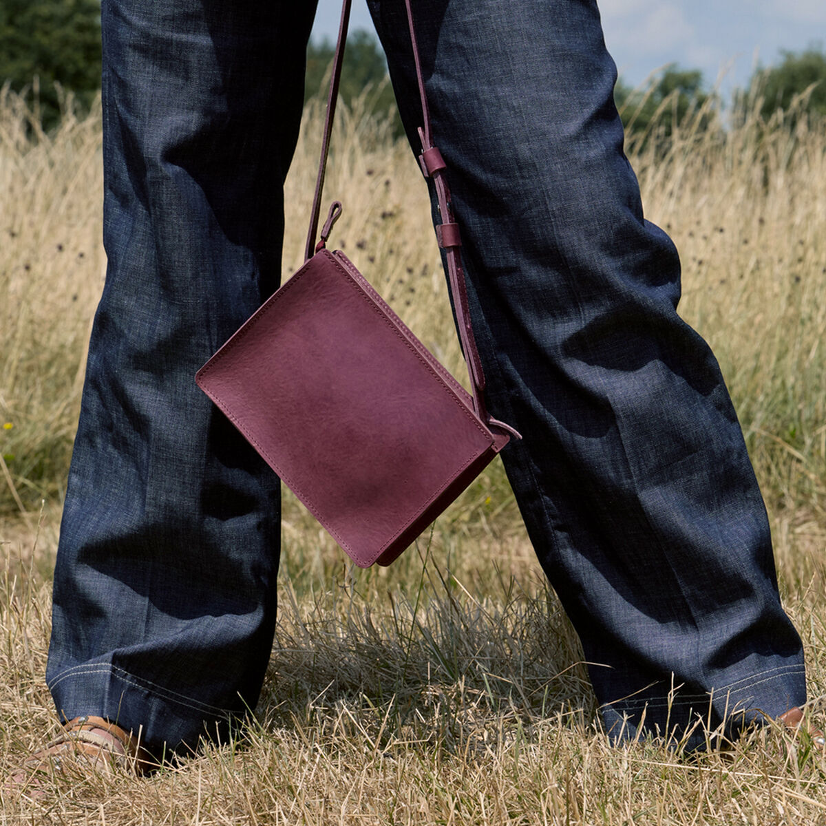 Eine bordeauxrote Kernleder-Umhängetasche mit schlichtem Design wird draußen in einer sommerlichen Wiese getragen. Zu sehen sind Beine in dunkelblauer Jeans, der Fokus liegt auf der Tasche, die lässig mit einem Schulterriemen getragen wird. Im Hintergrund wächst wildes Gras unter freiem Himmel.