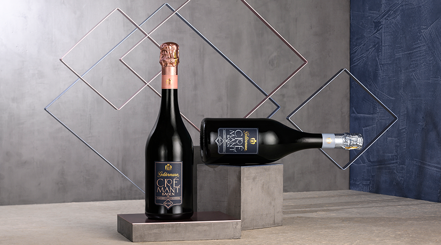 Zwei Flaschen Cremant Sekt, eine stehend mit roségoldener Folie, eine liegend mit silberner Folie, auf grauem Podest vor geometrischem Metallrahmen