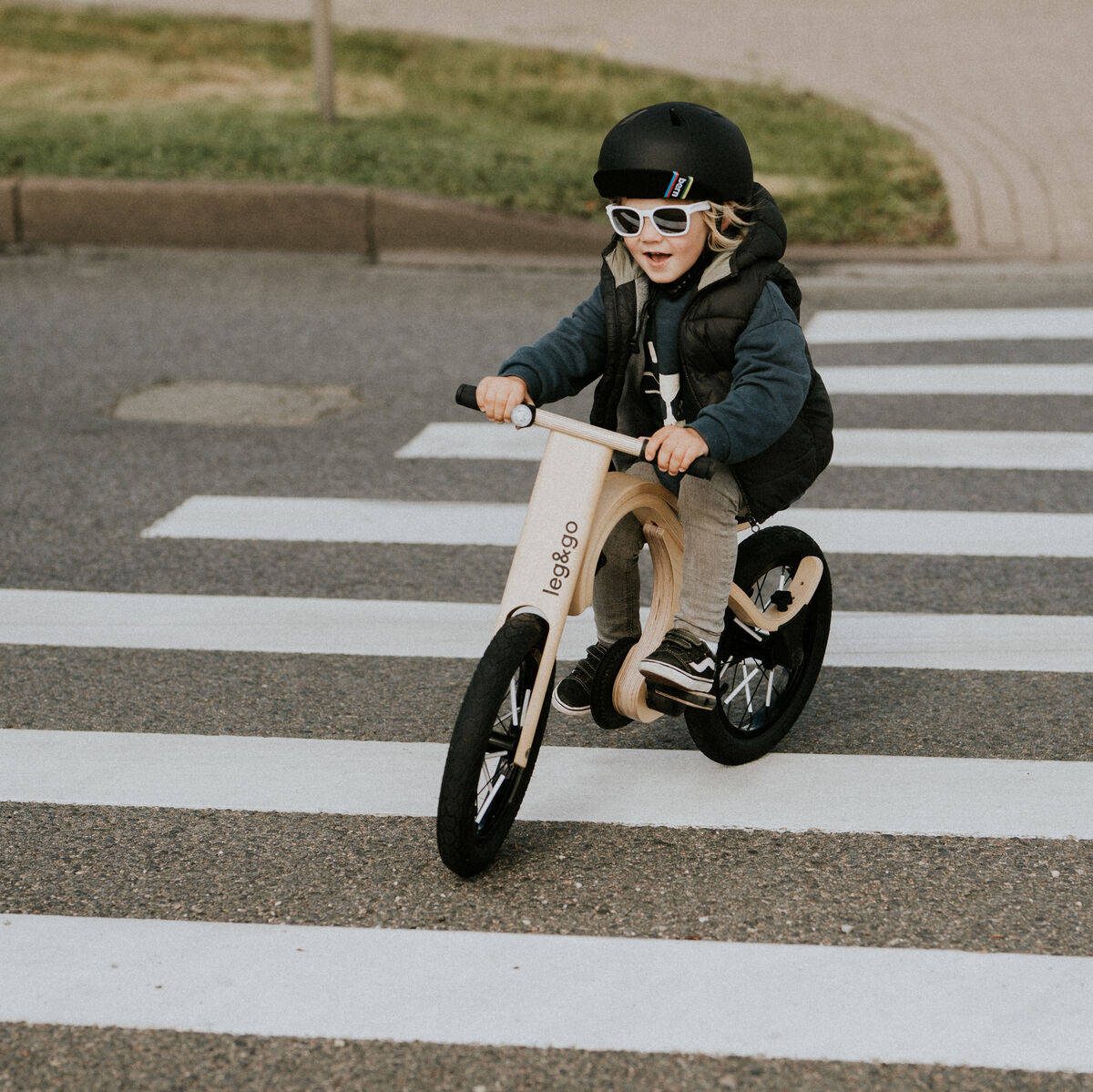 Ein Kind mit Helm und Sonnenbrille fährt auf einem hölzernen Laufrad mit Pedalerweiterung über einen Zebrastreifen. Das Kind ist draußen unterwegs, und das Bild zeigt einen sicheren Umgang mit dem Balance-Board-Zubehör für Fahrrad-Anfänger.