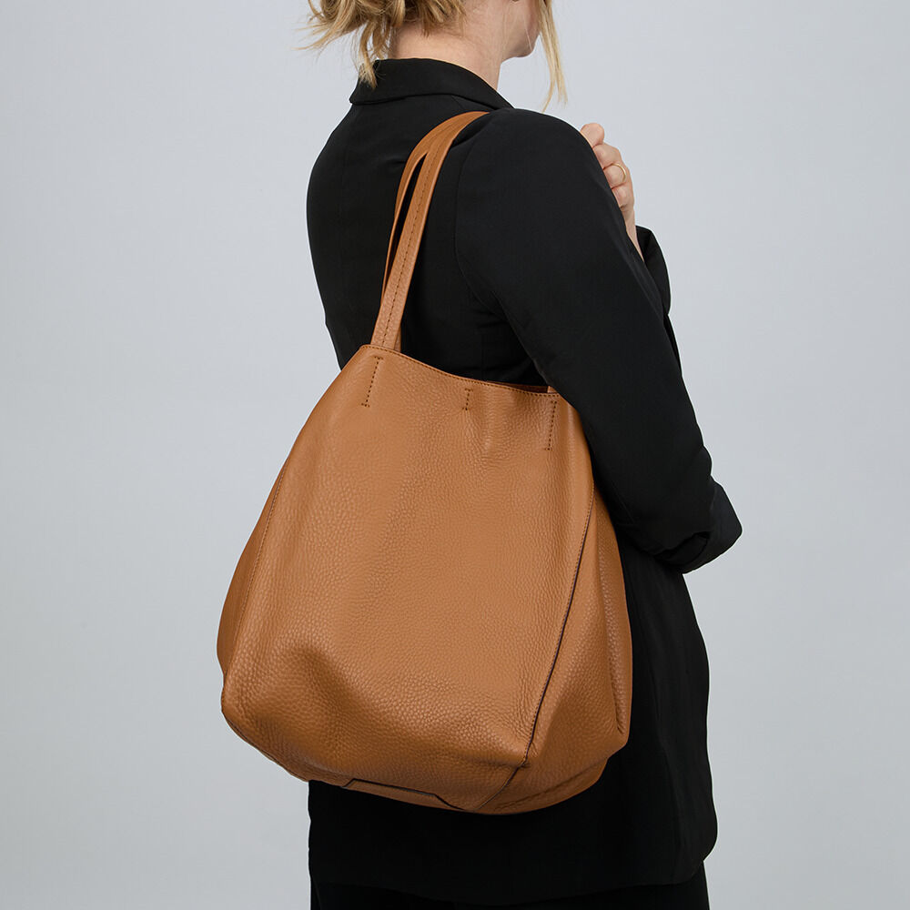 Frau im schwarzen Blazer trägt die ZEIT-Tasche »Cep« der Marke Guerer & Guerer in der Variante Cognac aus weichem, naturbelassenem Nappaleder. Die großzügig geschnittene Henkeltasche besitzt eine strukturierte Oberfläche und ein schlichtes, elegantes Design.
