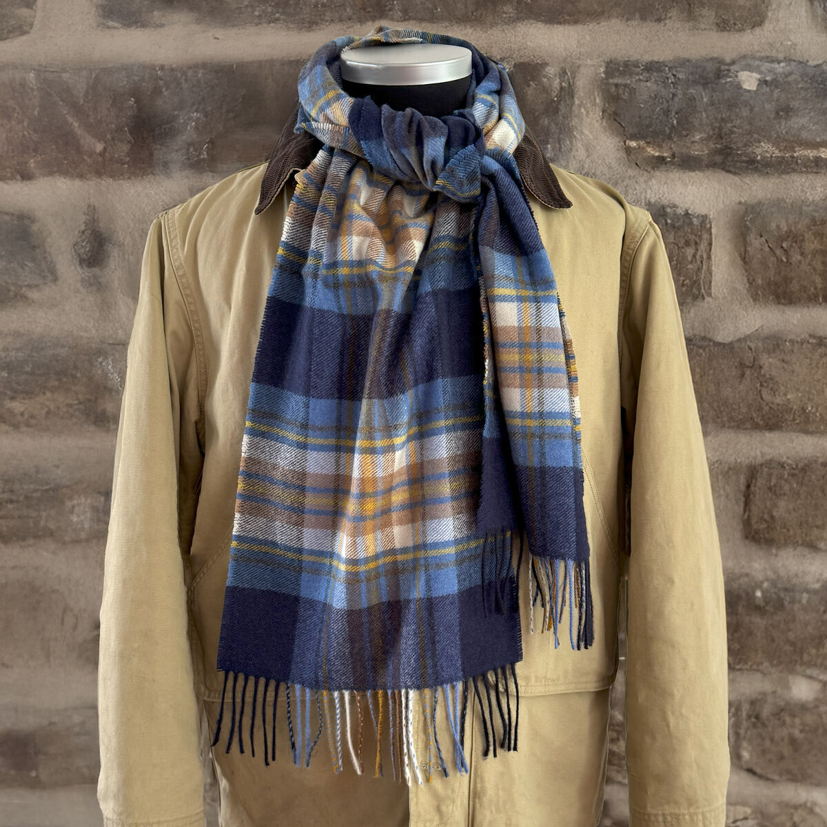 Ein eleganter Kaschmirschal mit blauem und beigem Karomuster ist um den Kragen einer beigen Jacke drapiert. Der Schal wirkt weich und luxuriös, ideal für Herbst- und Wintertage. Die Szene ist vor einer rustikalen Steinwand aufgenommen.