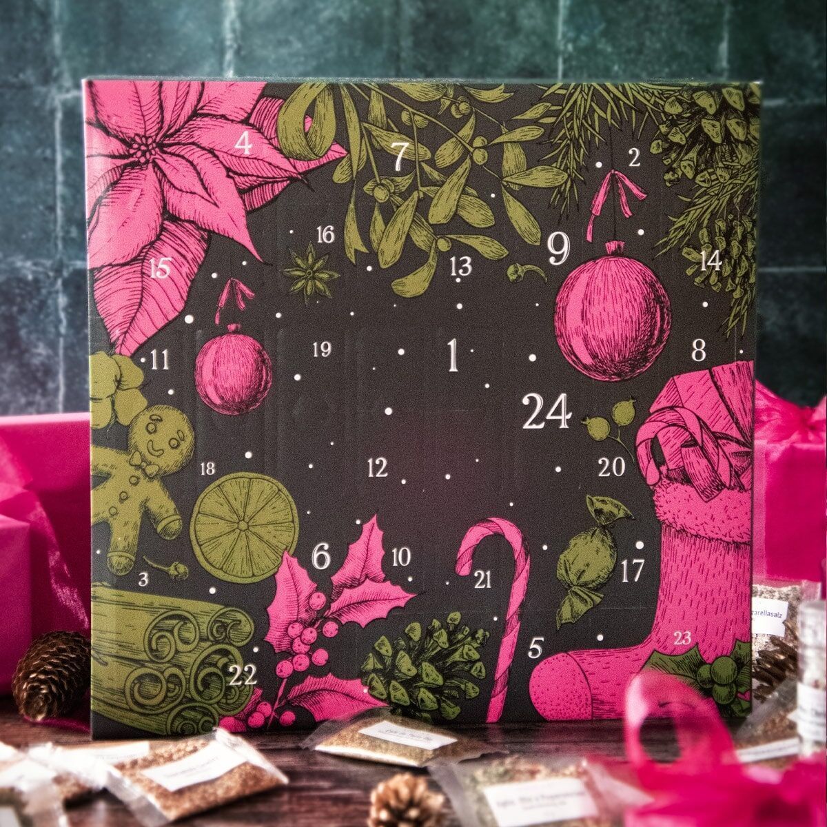 Ein Adventskalender mit roten und grünen Weihnachtsmotiven, wie Weihnachtsstern, Zuckerstange, Handschuh und Christbaumkugeln, auf schwarzem Hintergrund. Es gibt nummerierte Türchen von 1 bis 24. Im Hintergrund und Vordergrund sind zylindrische Glasbehälter mit losen Teeblättern zu sehen, umgeben von rosa Geschenkpapier und kleinen Kiefernzapfen.