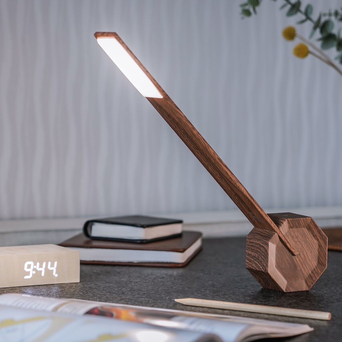 Die Octagon Tischlampe »One« aus Holz steht in einem modernen Büro. Neben der Lampe sind Bücher, ein Smart-Home-Wecker und ein Stift zu sehen. Die Lampe hat eine minimalistische und moderne Bauform und leuchtet hell.