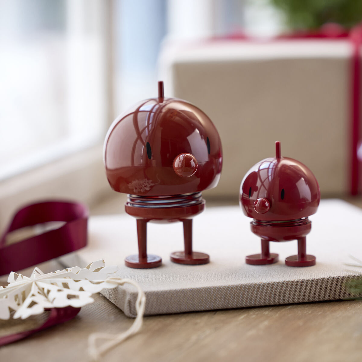Zwei Hoptimist Bumble Maroon Figuren der Marke Hoptimist in glänzendem Bordeauxrot stehen auf einem weißen Geschenk, dahinter weitere Geschenke mit rosa Schleifen; Dekorationselemente sorgen für festliche Atmosphäre.