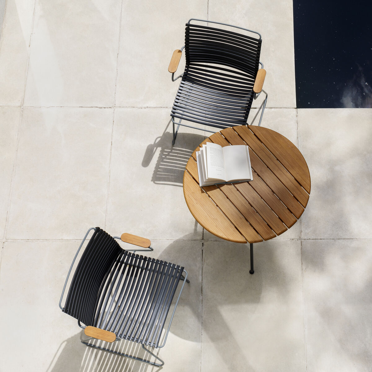 Zwei »CLICK Dining Chair« mit Armlehne und ein runder Holztisch auf weißem Terrassenboden neben einem dunkel reflektierenden Wasserbecken. Auf dem Tisch liegt ein aufgeschlagenes Buch.