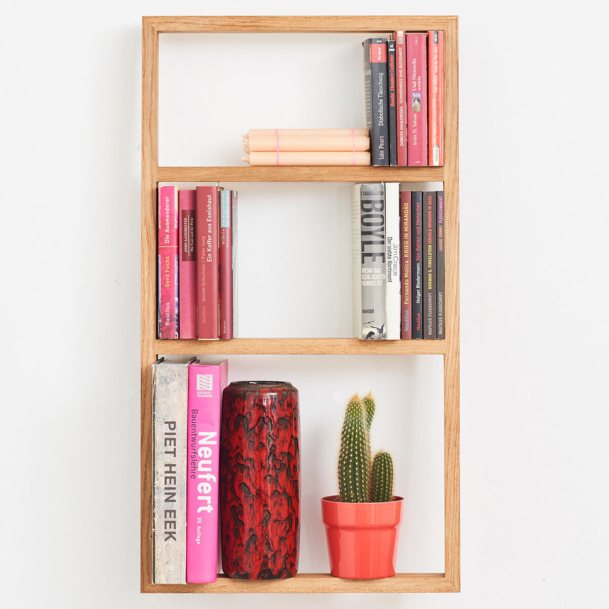 Ein minimalistisches Wandregal aus Holz, gefüllt mit Büchern und Dekorationsgegenständen, einschließlich einer roten Vase und einem Kaktus in einem roten Übertopf.