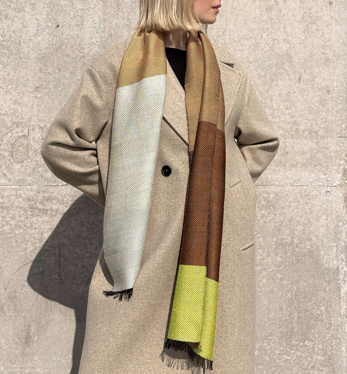 Frau mit blondem Bob trägt einen Seidenschal in Naturtönen wie Beige, Braun und Hellgelb über einem beigen Mantel vor grauer Betonwand. Das Outfit wirkt elegant und zeitlos.