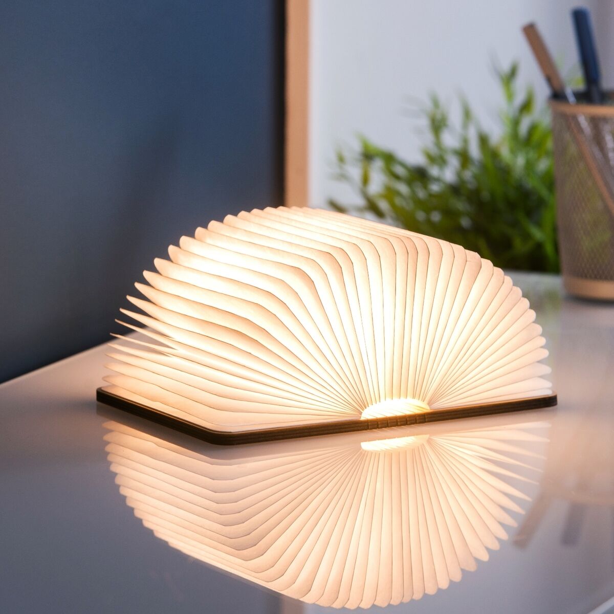 Moderne, aufklappbare Buchlampe mit walnussfarbenem Holzdeckel. Perfekt als stimmungsvolle Beleuchtung auf einem weißen Tisch, mit einem Topf Stifte und einer Pflanze im Hintergrund.