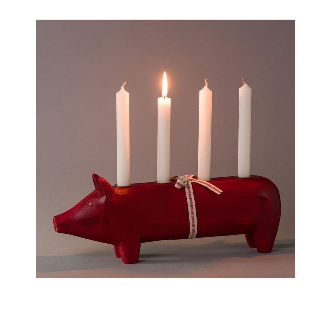 Kerzenhalter »Holz-Schwein« der Marke Maileg aus rotem Holz mit vier weißen Stabkerzen, wovon eine brennt. Das Schwein steht auf einem Tisch vor neutralem Hintergrund, einfache Details, dekorativ und modern, 38 cm lang, mit weißem Band um den Körper.