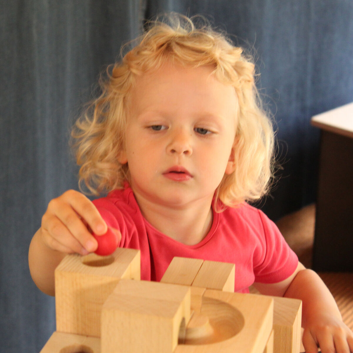 Ein kleines Kind spielt mit einer hölzernen Kugelbahn auf einem Tisch. Das Spielzeug besteht aus naturfarbenen Holzblöcken. Das Kind trägt ein pinkfarbenes T-Shirt.