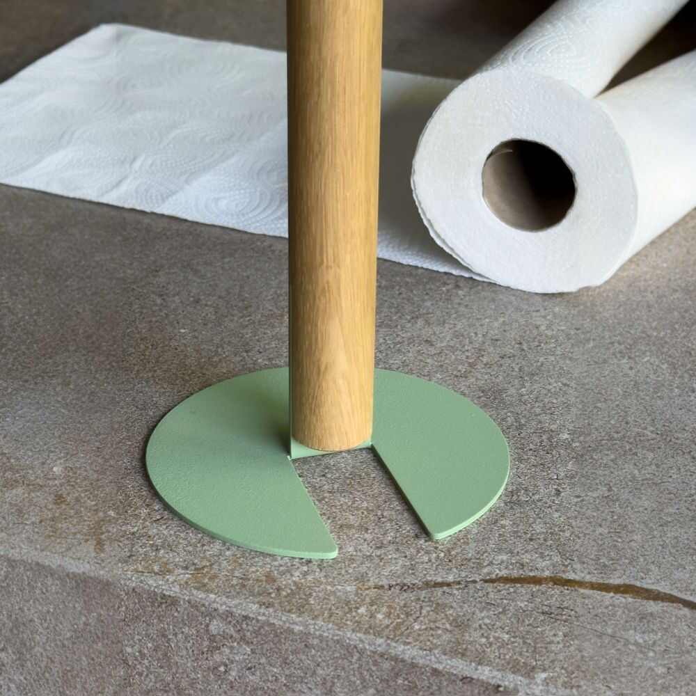 Nahaufnahme eines Küchenrollenhalters auf einer grauen, rauen Stein-Arbeitsplatte: Ein senkrechter Stab aus hellem Naturholz steckt in einem flachen, salbeigrünen, kreisförmigen Metallfuß mit einer offenen Aussparung. Dahinter liegen eine teilweise abgerollte und eine aufgerollte weiße Küchenpapierrolle.