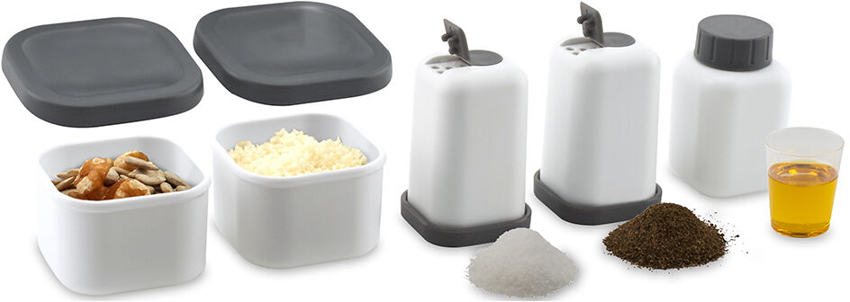 Verschiedene Behälter und Dosen eines Lunch Care Systems, inklusive zwei quadratische graue Deckel, zwei weiße Behälter mit geriebenem Käse und Nüssen, drei weiße Flaschen mit grauen Deckeln und Stöpseln, eine mit weißen, eine mit schwarzen und eine mit braunen Pulver gefüllt, und ein kleiner transparenter Becher mit gelber Flüssigkeit.