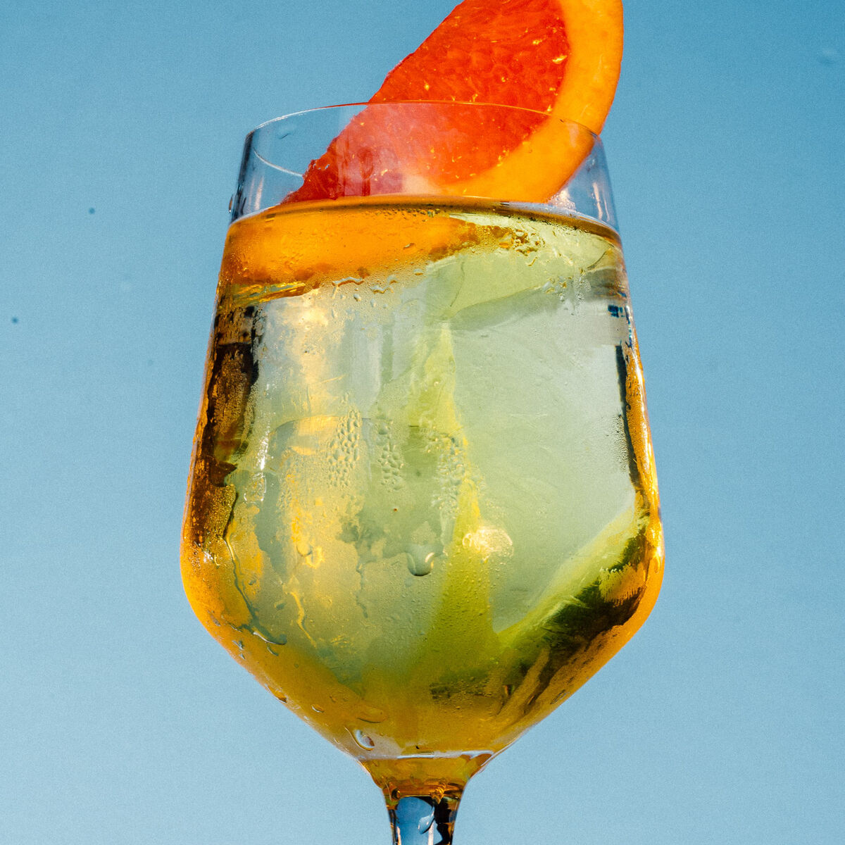 Ein Aperitif »Bright Lemon« in einem Weinglas mit Eiswürfeln und einer Orangenscheibe vor einem blauen Hintergrund.