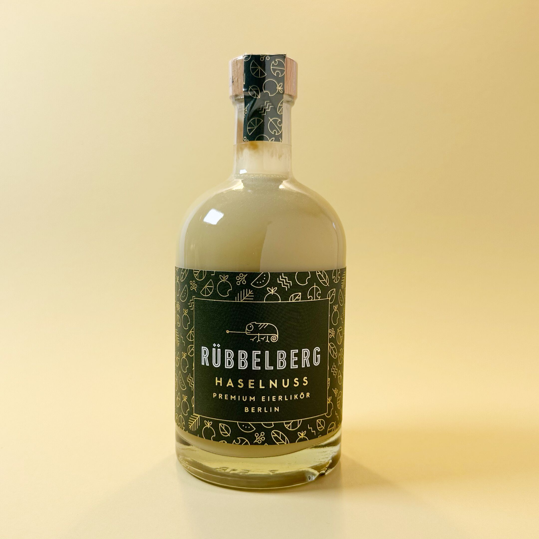 Eine stilvoll fotografierte Flasche Premium Eierlikör Haselnuss der Marke Rübbelberg steht vor einem einfarbig beigen Hintergrund. Das Etikett ist dunkelgrün mit goldenen und weißen Details gestaltet und zeigt deutlich den Produktnamen sowie Haselnuss-Illustrationen. Die Flasche ist verschlossen und elegant präsentiert.