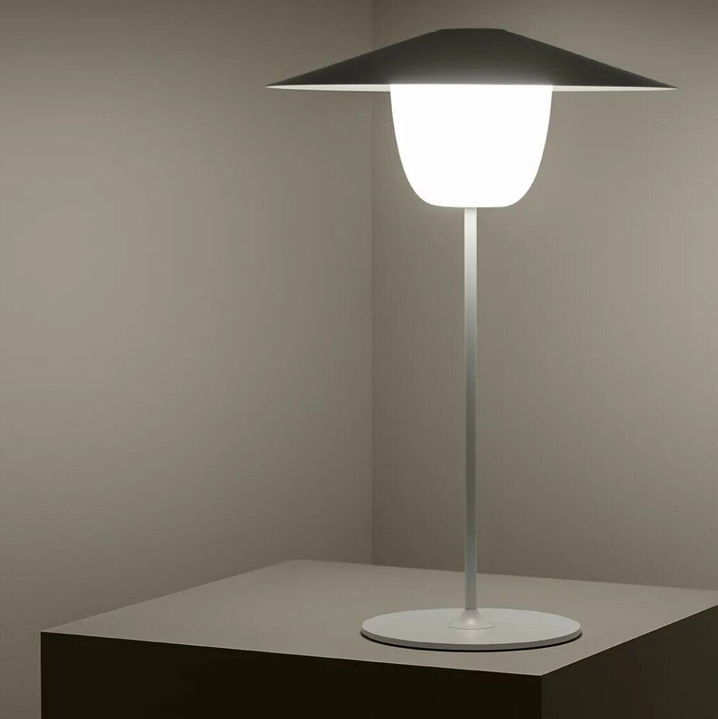 Eine moderne, mobile LED-Tischleuchte mit schlichtem Design steht auf einem kubischen Tisch in einem minimalistisch eingerichteten Raum. Die Leuchte strahlt ein weiches, weißes Licht aus.