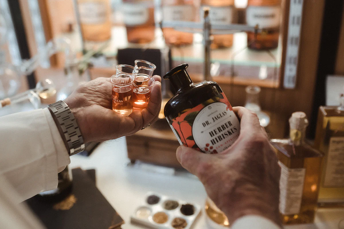Person hält drei kleine Gläser mit bernsteinfarbener Flüssigkeit und eine Flasche von Dr. Jaglas in einer Bar oder einem Geschäft