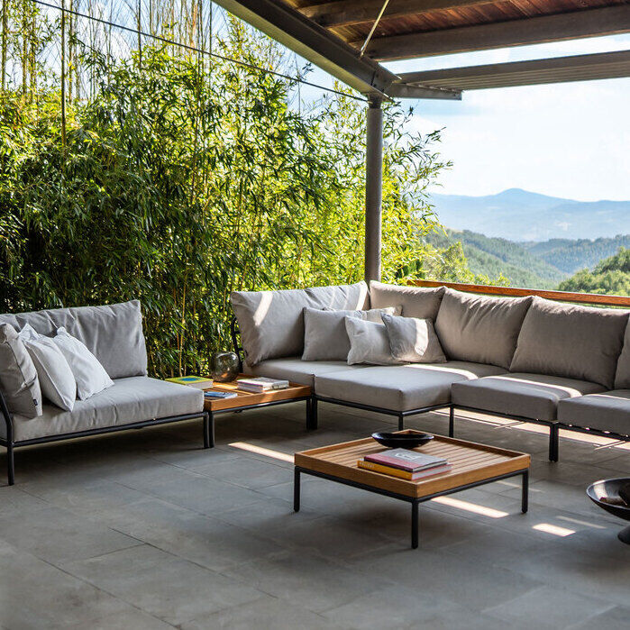 Ein modernes, graues Ecksofa der Lounge LEVEL 2 Serie steht in einer überdachten Terrasse mit Blick auf eine grüne, bergige Landschaft. Im Vordergrund befinden sich zwei niedrige Holztische. Die Umgebung ist mit Bambuspflanzen und anderen Grüntönen dekoriert.