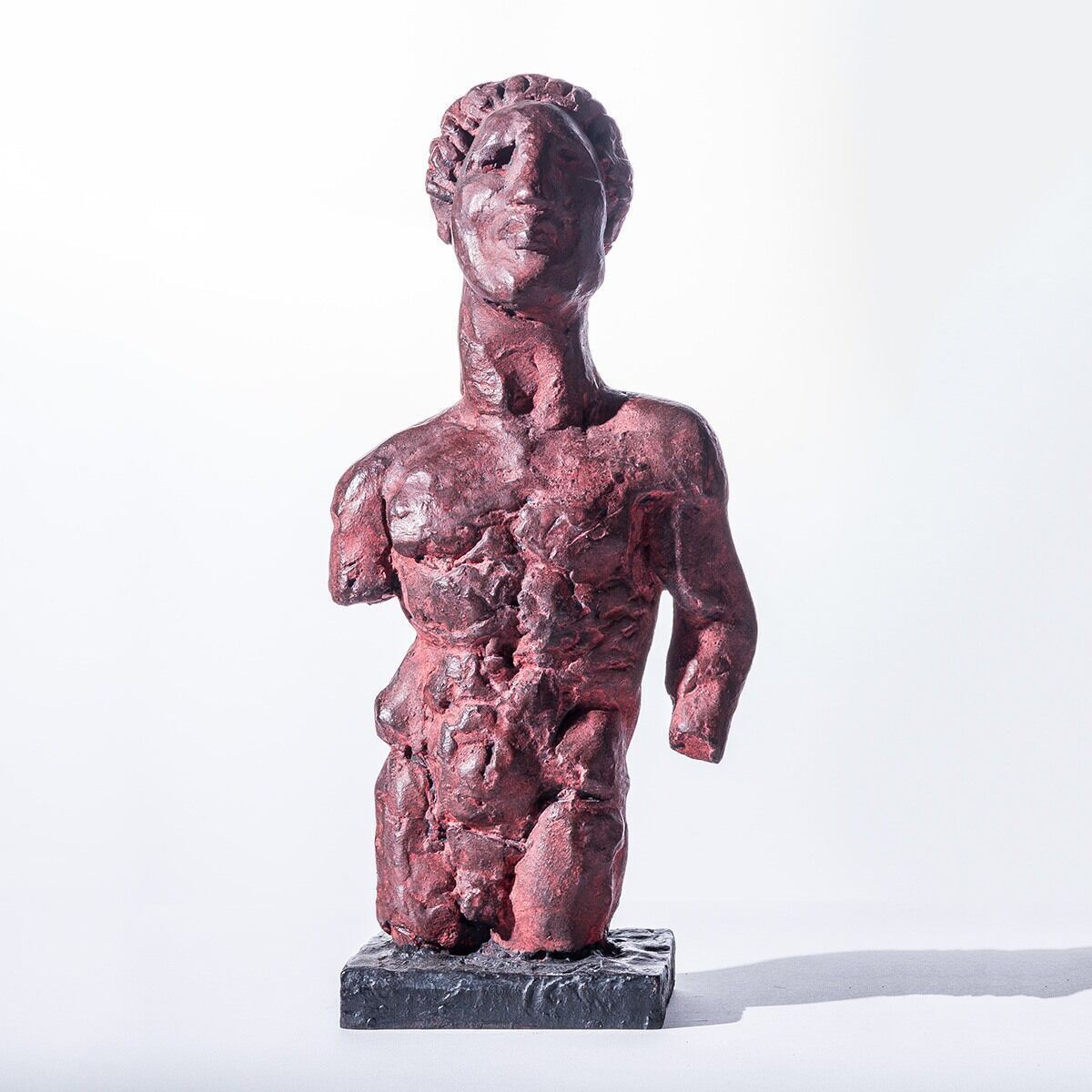 Die Skulptur »Das Grundgesetz«, 2013 von Markus Lüpertz ist eine abstrakte, bronzefarbene Figur, die teilweise in roten und schwarzen Tönen gehalten ist. Sie zeigt einen menschlichen Oberkörper und Kopf, wobei detaillierte Strukturen und Oberflächen zu erkennen sind. Die Figur steht auf einem grauen Sockel und wird von einer diffusen, weißlichen Hintergrundbeleuchtung hervorgehoben.