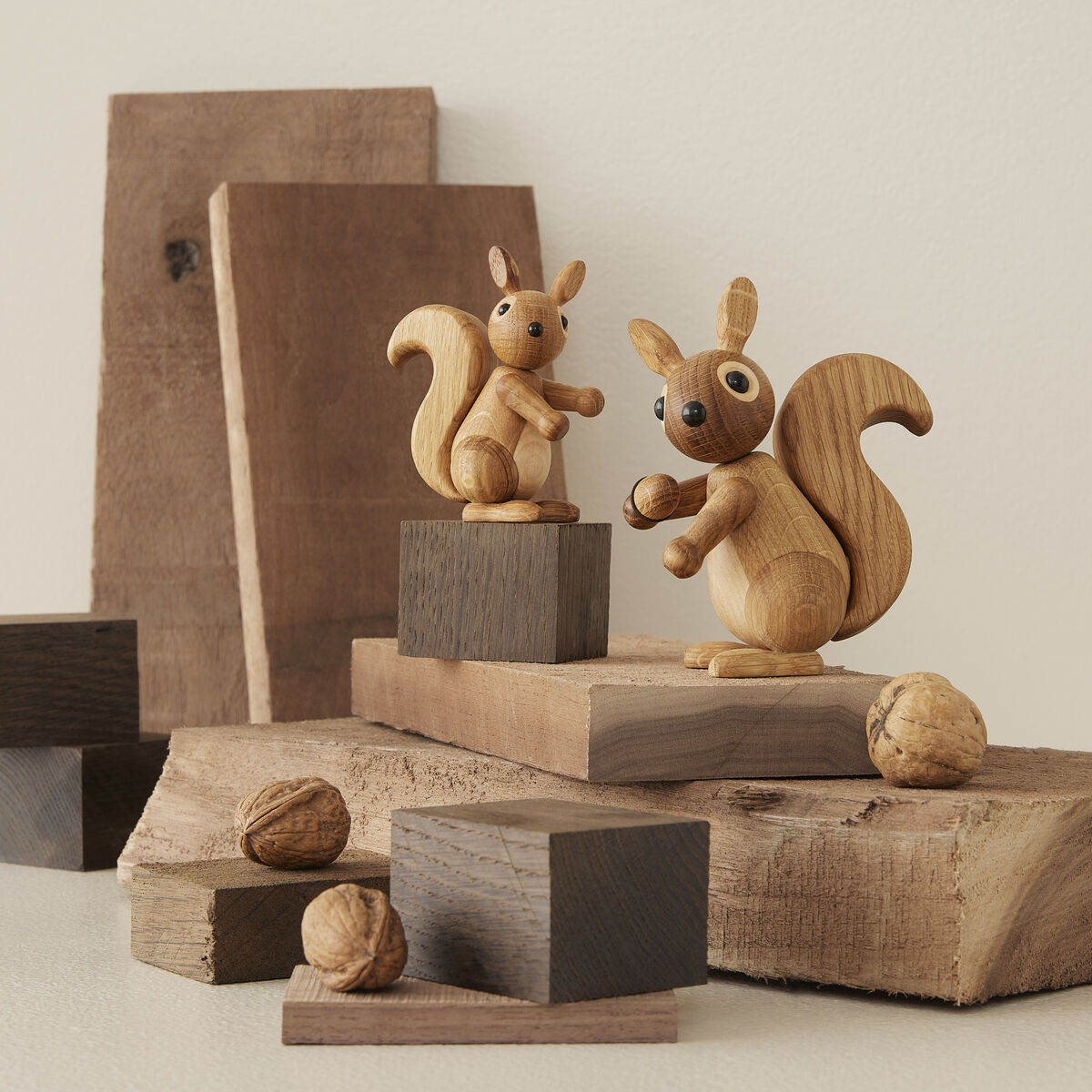 Zwei niedliche Eichhörnchen-Holzfiguren namens Hazel stehen auf verschieden großen Holzstücken, umgeben von Walnüssen. Die Szene wirkt natürlich und minimalistisch und eignet sich ideal als Dekoration für Wohnräume oder Kinderzimmer.